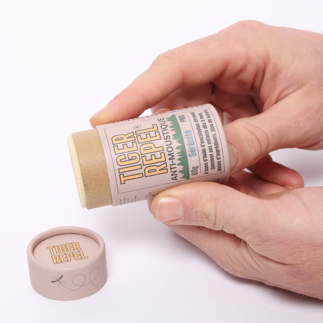 Une main tenant un stick anti-moustique Tiger Repel "Sérénité" ouvert pour montrer son application facile. Le stick est contenu dans un tube en carton biodégradable avec son couvercle posé à côté sur un fond blanc.